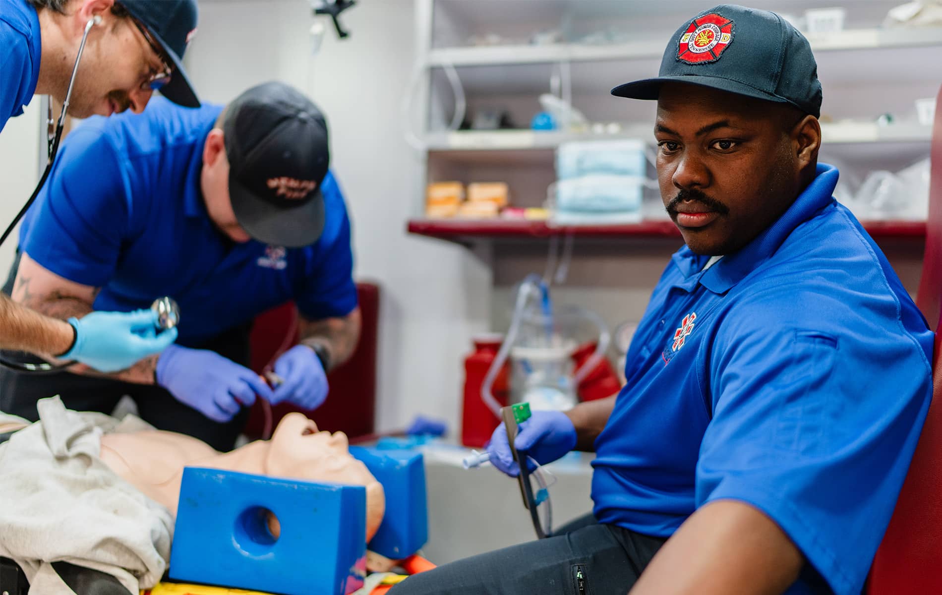 Three emergency medical responders in blue uniforms practice airway management on a medical training mannequin in a clinical lab setting, with equipment and supplies visible in the background.