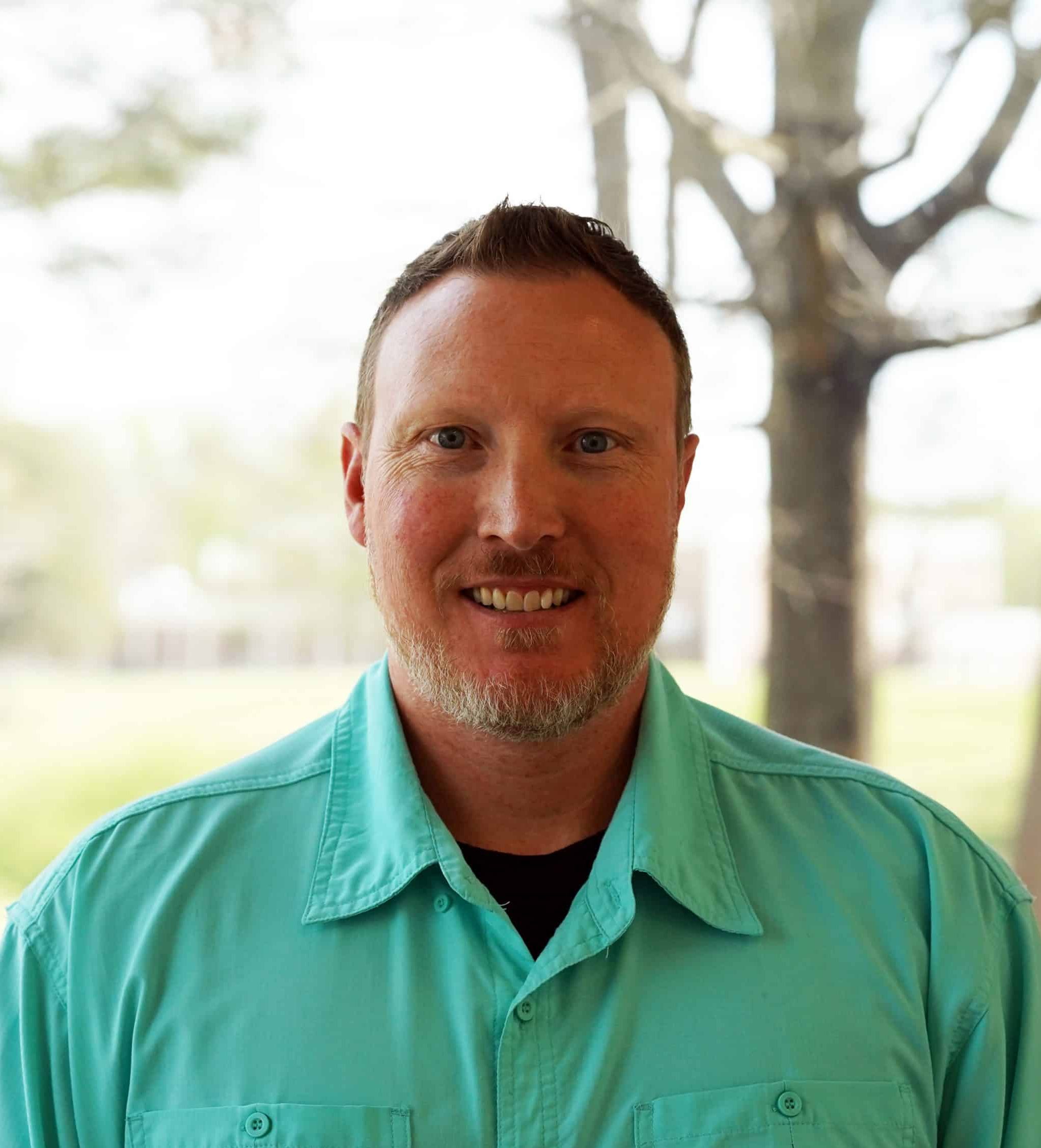 Photo of Mr. Clay Boyd standing in front of a glass window, with the campus in the background, blurred. Clay is wearing a green button shirt and smiling toward the camera.