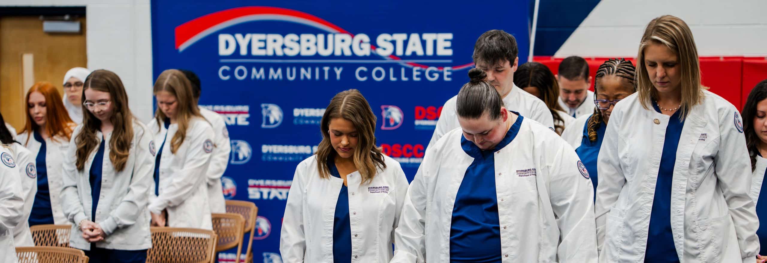 A group of Dyersburg State Community College nursing students stand together in white clinical jackets and blue scrubs during the nursing pinning ceremony. Many students have their heads bowed in a moment of reflection or prayer. A large DSCC-branded backdrop featuring the college logo is displayed behind them inside a gymnasium or event space. Chairs are arranged in the foreground, and the atmosphere appears solemn and respectful.