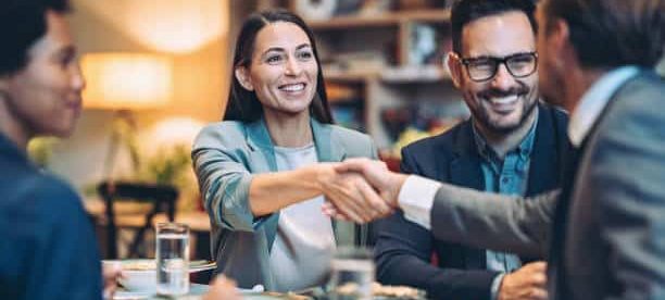 Four professionals seated around a table during a Lunch & Learn event smile and shake hands while enjoying a meal and conversation. Glasses of water, plates of food, and a relaxed meeting atmosphere are visible in the warmly lit setting.