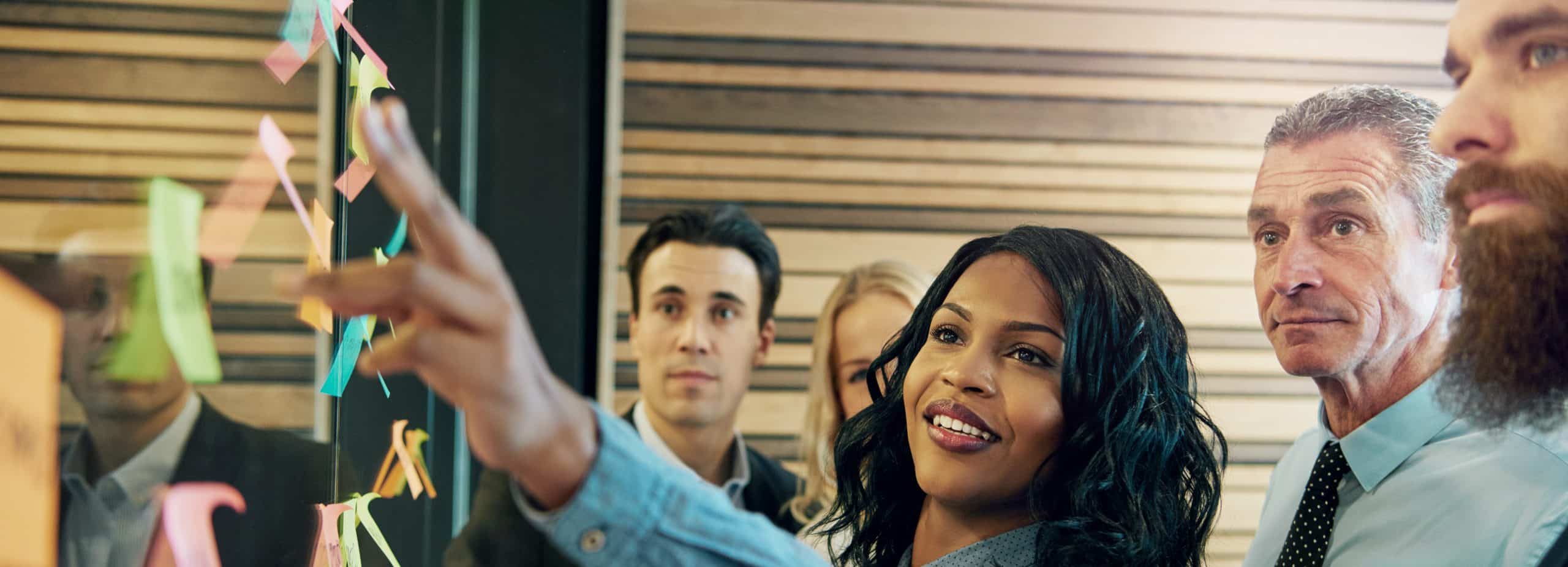 Image of a diverse group of business professionals collaborating with a leader. There are sticky notes on the wall, and the leader is explaining the process.