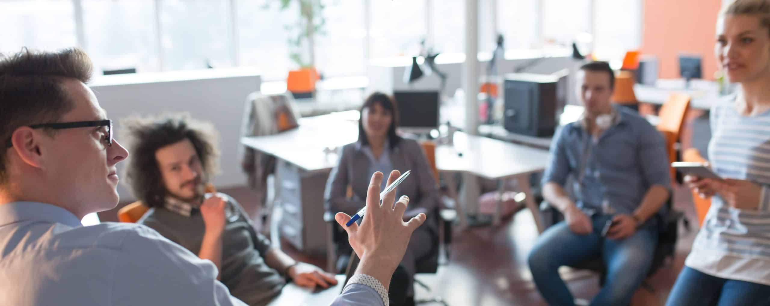 Five people gathered in a modern office for a team discussion. One person in the foreground gestures with a pen while seated participants listen and engage in conversation. Desks, computers, and office furnishings are visible in the background.