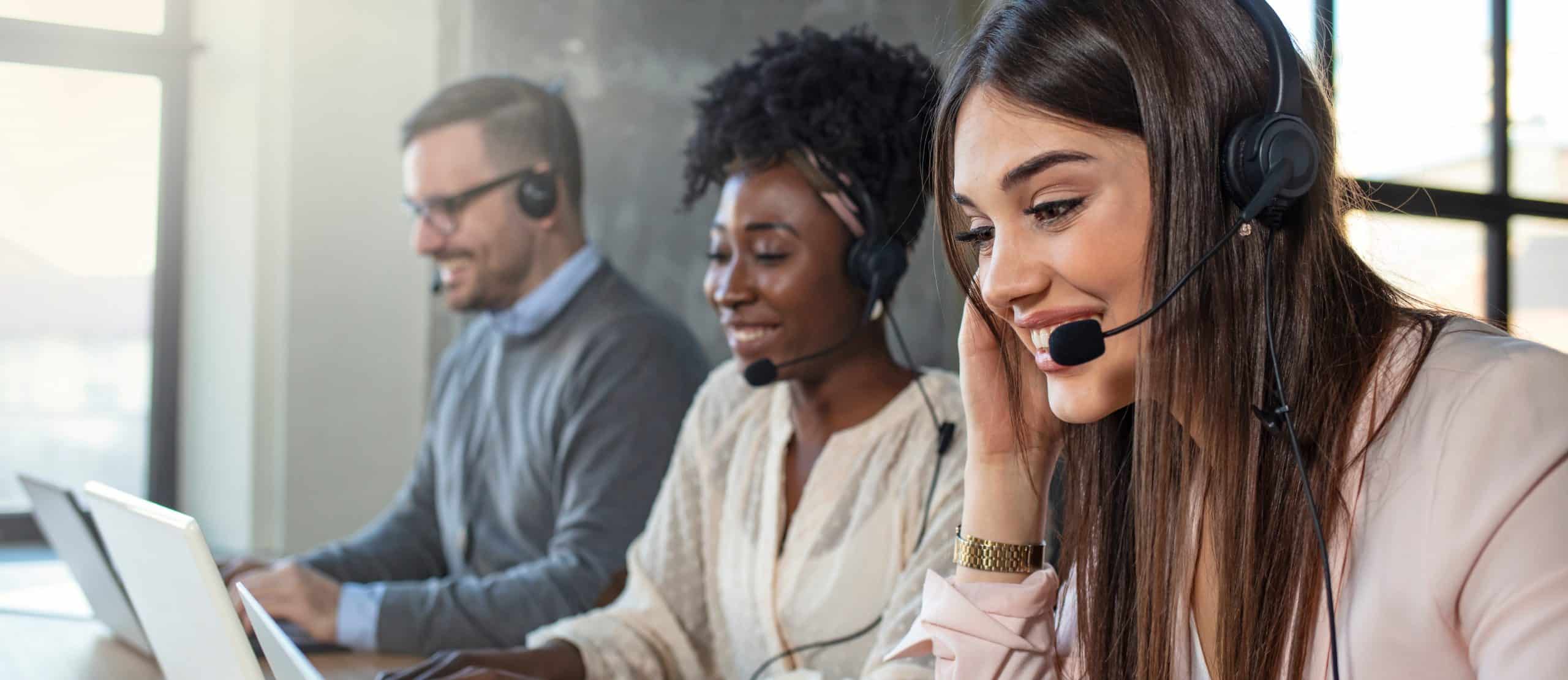 Three call center agents seated at desks wearing headsets and using computers in a modern office. The image depicts customer support, help desk services, and communication with clients through phone and online channels.