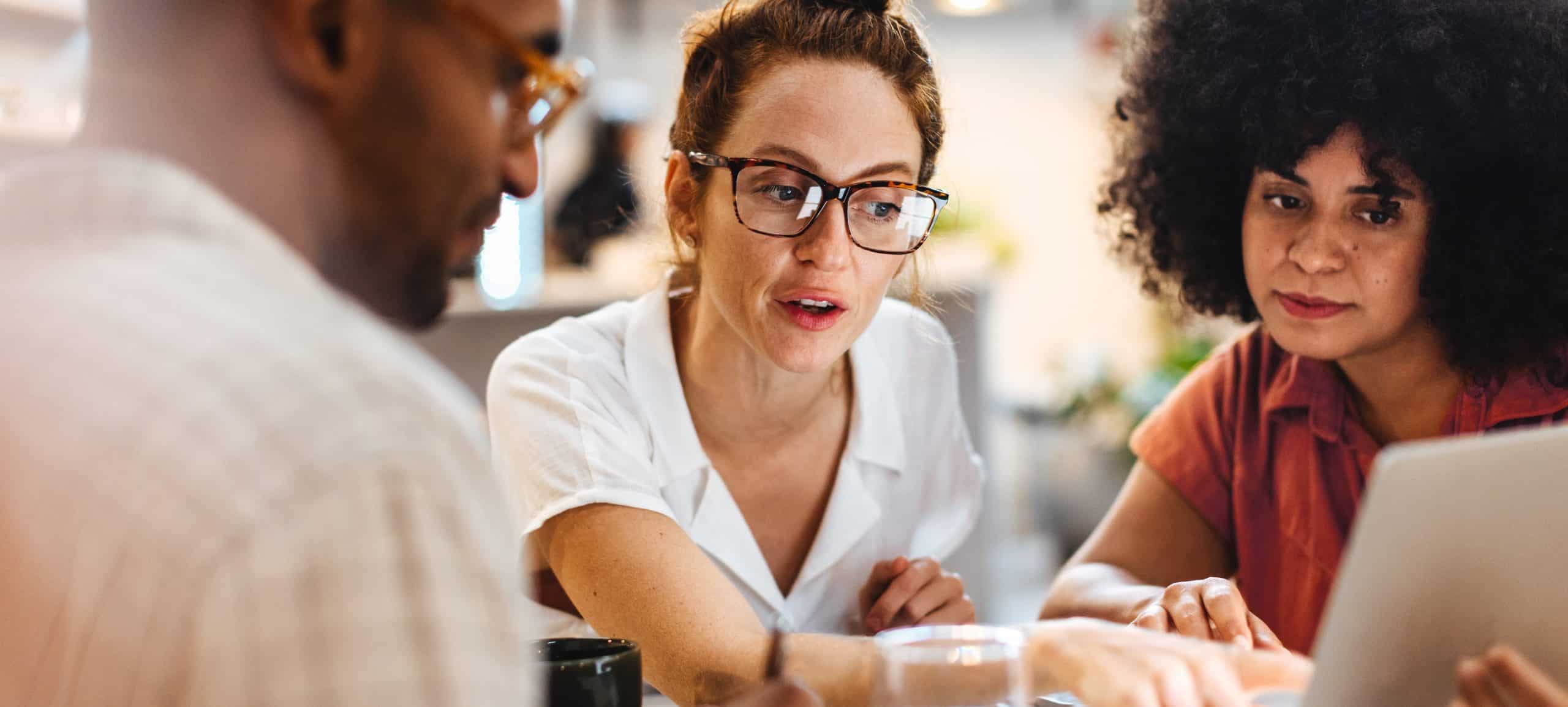 Three business professionals sit at a table with drinks and a laptop in a business meeting.