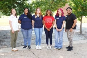 Six people stand together in a line on a college campus pathway with trees providing shade in the background. One person wears a red DSCC T-shirt, while others wear DSCC-branded polo shirts and name badges. The group is photographed outdoors near campus seating and landscaping.