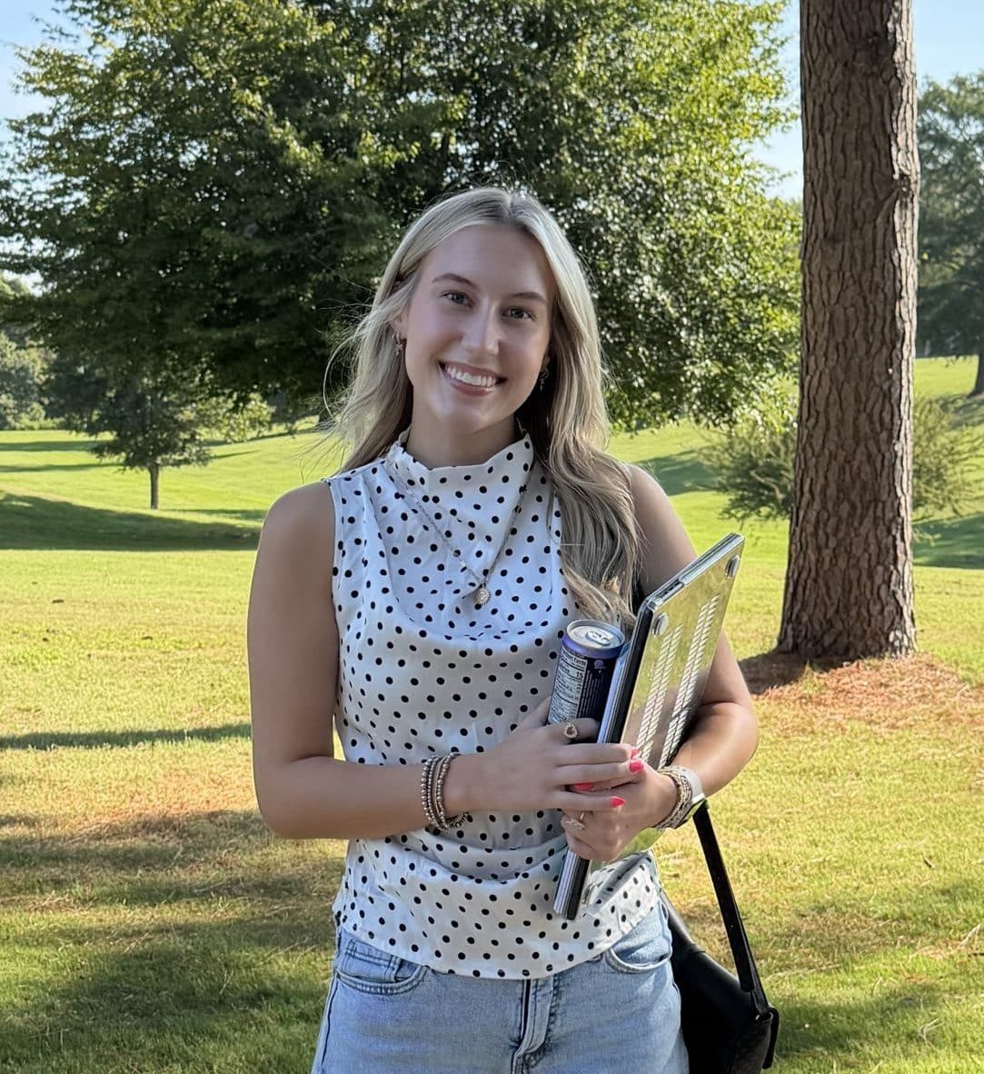 A student stands outdoors on a campus walkway holding a notebook, binder, and drink. The student is wearing a sleeveless white polka-dot top, light blue jeans, and white sandals, with a shoulder bag. Behind the student is a large grassy lawn, mature trees, and a campus building visible in the distance. The photo was taken on a sunny day with clear blue skies and natural lighting.