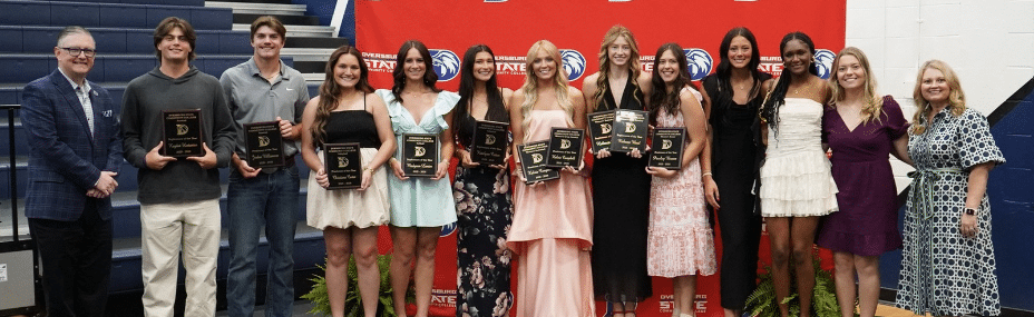 Group of DSCC student-athletes standing in front of a red DSCC backdrop and posing with athletic awards.