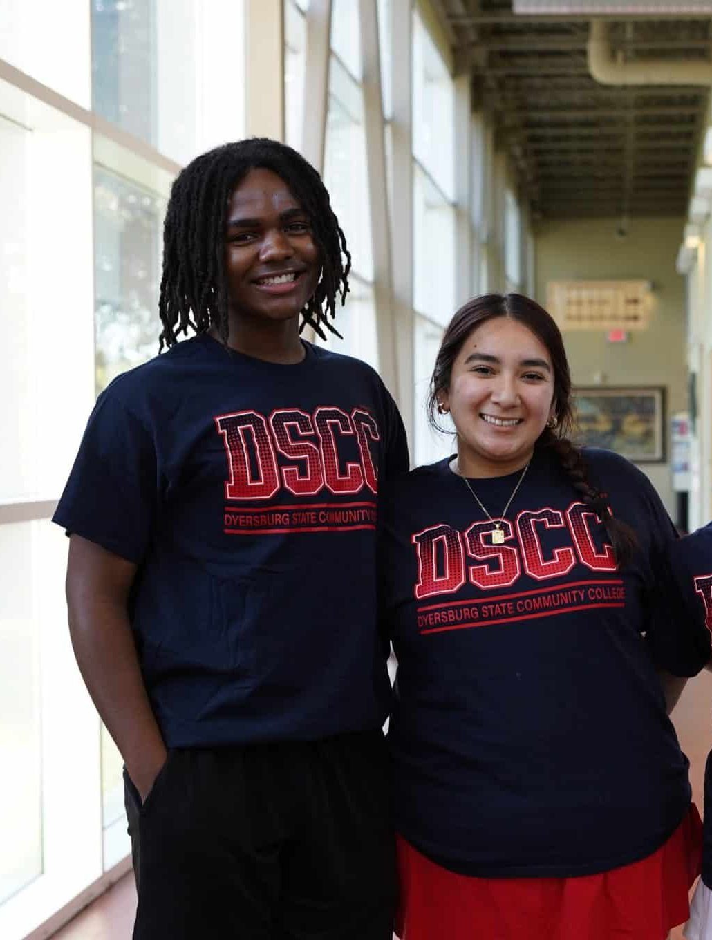 Two students smiling and posing in a well-lit hallway.