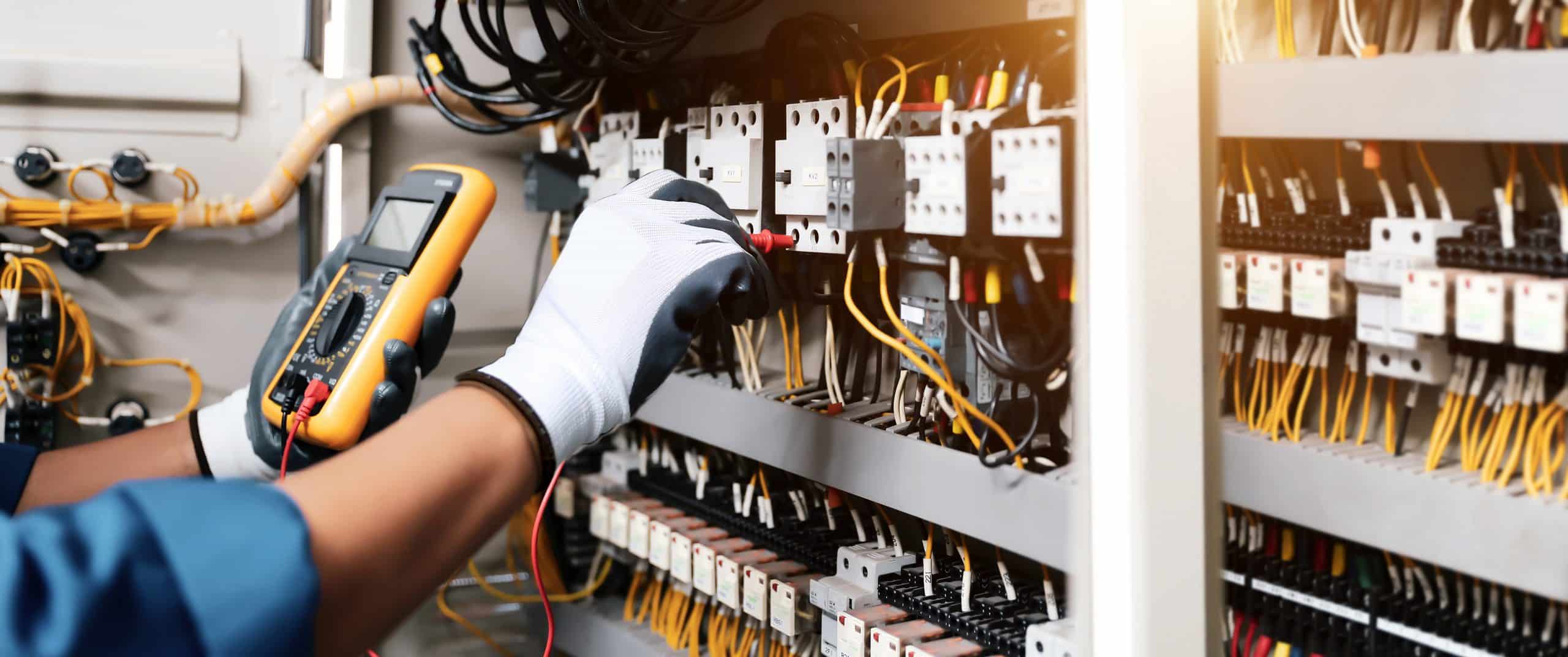 A gloved electrician uses a yellow digital multimeter to test connections inside an open electrical control panel filled with circuit breakers, relays, and yellow and white wiring.