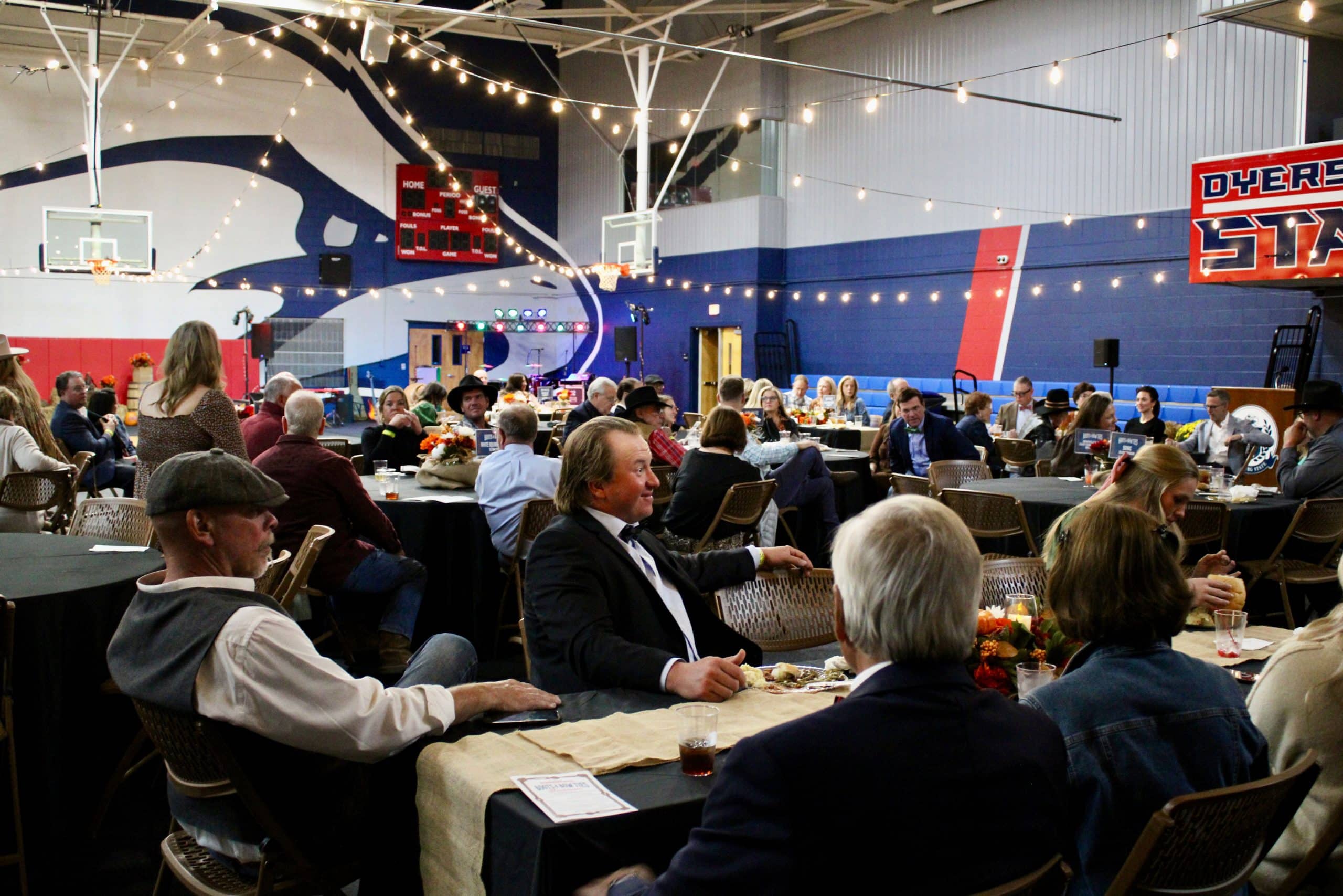 Image of community members sitting at tables inside the gymnasium with low lighting and hanging lights.